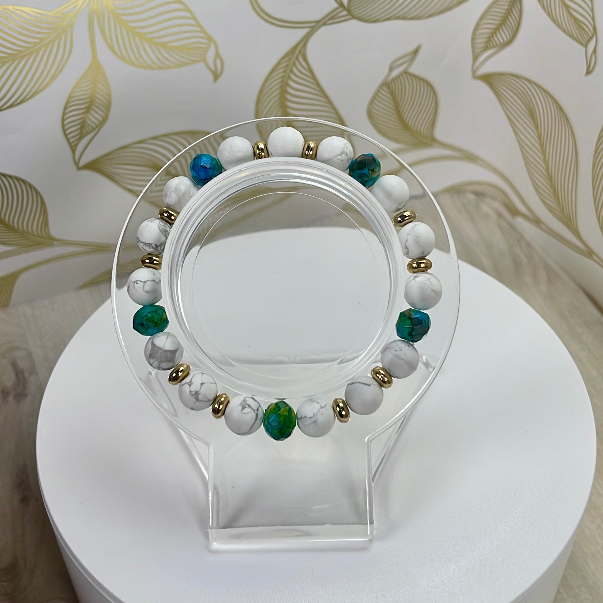 A soft, matte white bracelet howlite paired with blue-green rondelle beads and gold details on a clear stand with a decorative background