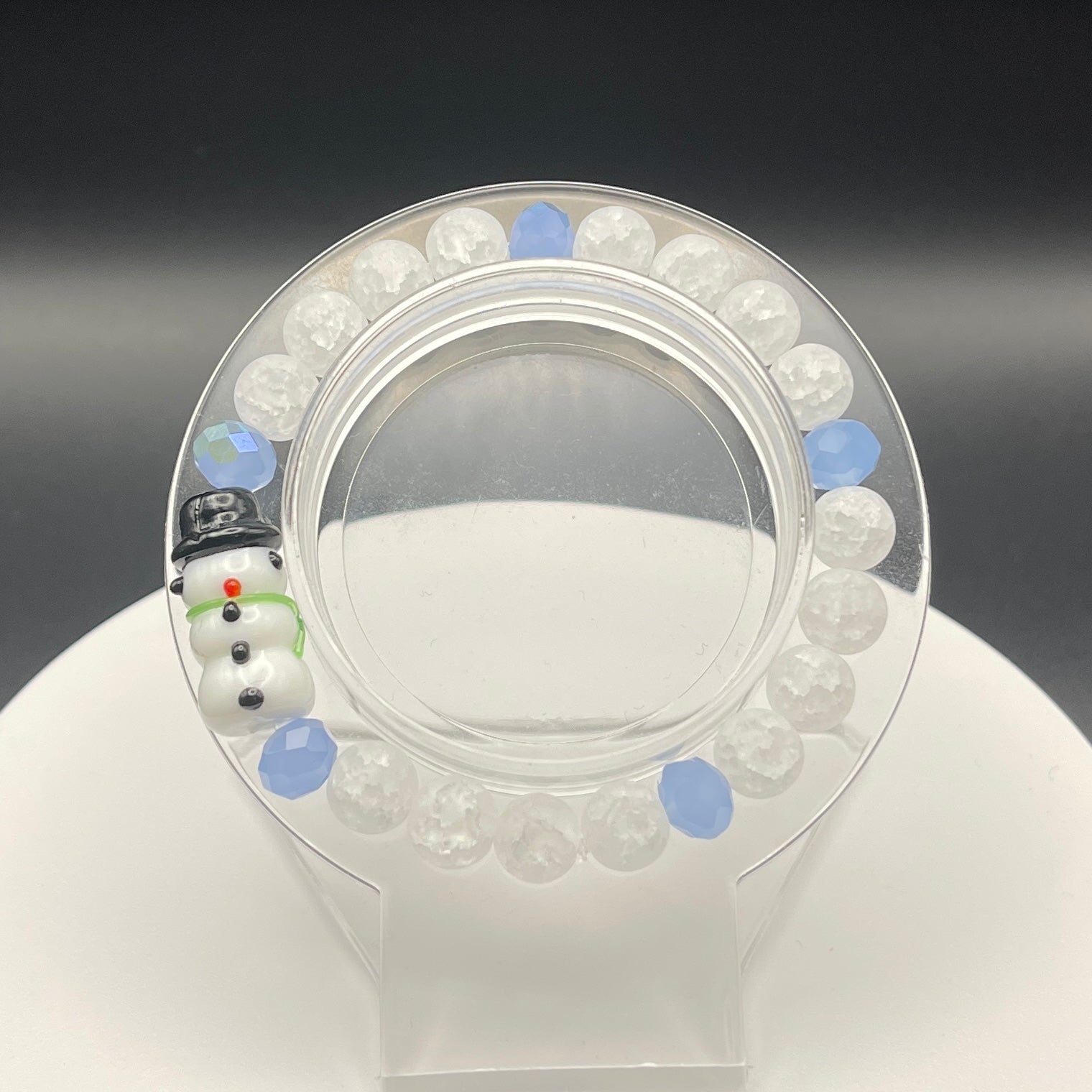 Clear plastic bracelet display with white and blue beads and a snowman-shaped bead on a black background
